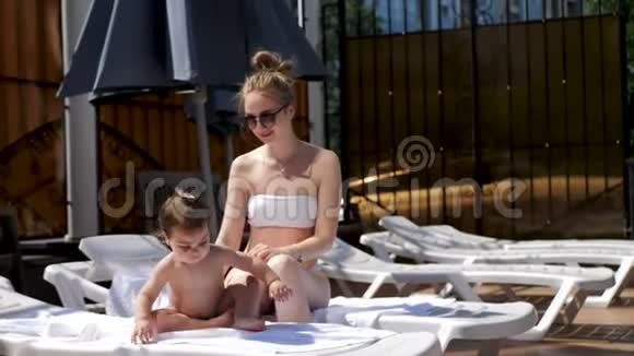 暑假里妈妈和小女儿坐在开放的游泳池里的躺椅上视频的预览图