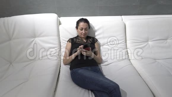 一个带着手机的可爱女人坐在白色沙发上穿着便服她在说话看着什么视频的预览图