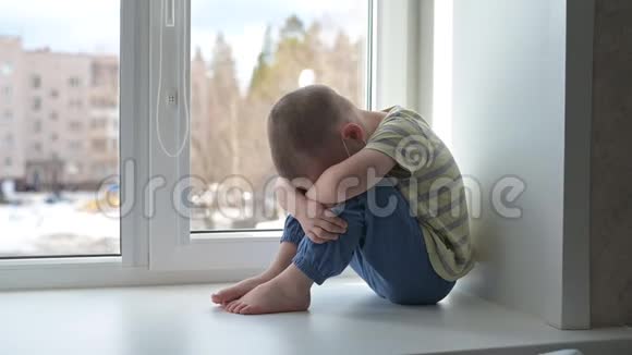 悲伤的孩子戴着防护口罩孤独地坐在窗户附近检疫呆在家里慢动作手持视频的预览图