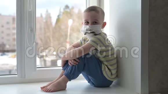 悲伤的孩子戴着防护口罩孤独地坐在窗户附近检疫呆在家里慢动作手持视频的预览图
