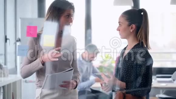 两个年轻的商业女性一起工作在墙上的玻璃并张贴它的贴纸在合作空间视频的预览图