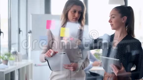 两个年轻的商业女性一起工作在墙上的玻璃并张贴它的贴纸在合作空间视频的预览图