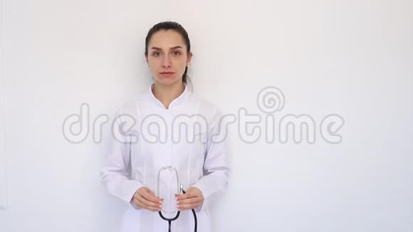 年轻的微笑医生女女孩穿着白色的医疗外套把听诊器放在耳朵里直接看着相机视频的预览图