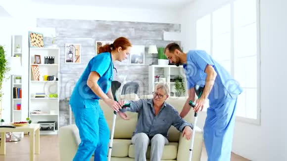 女医生和她的助手用拐杖帮助老妇人从沙发上站起来视频的预览图