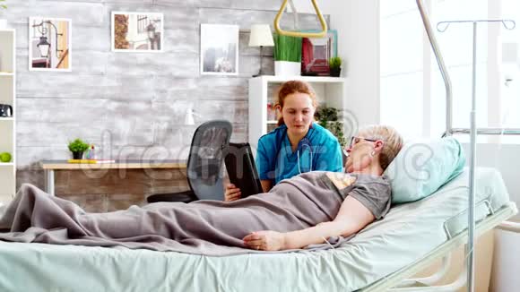 女性照顾者帮助躺在病床上的老年残疾妇女使用数码平板电脑视频的预览图