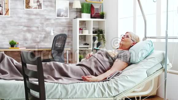 在明亮的养老院里躺在病床上的老太太和一位女性照顾者交谈视频的预览图