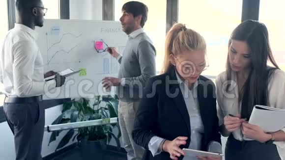 两位优雅的年轻商务女性在数码平板电脑前工作而她的同事则在C语言中的白板前讲解这个项目视频的预览图