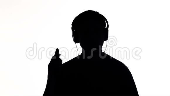 一个年轻快乐的女人戴着大耳机听音乐拿着智能手机的剪影女孩在白色的隔离巴上跳舞视频的预览图