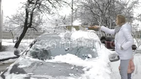 一个年轻女孩在雪后用刷子清洗覆盖着雪的灰色汽车视频的预览图