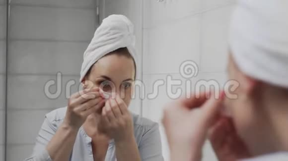 头上戴毛巾的成年女性将胶原蛋白垫放在眼睛下面看着镜子视频的预览图