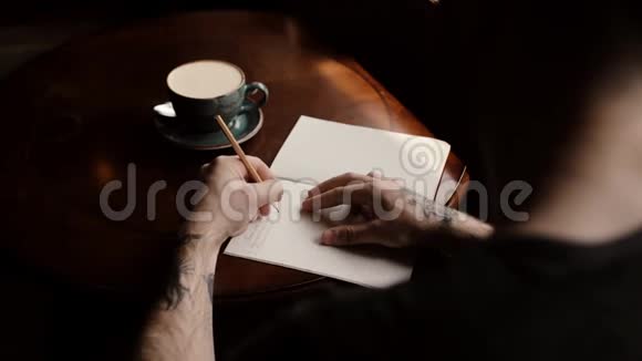 男人纹身的手在咖啡馆特写的咖啡桌上的笔记本上写字视频的预览图