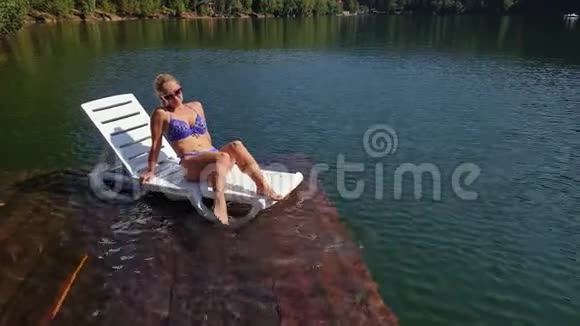 女人戴着太阳镜和泳衣躺在日光浴床上女孩在洪水木水下码头休息路面是视频的预览图