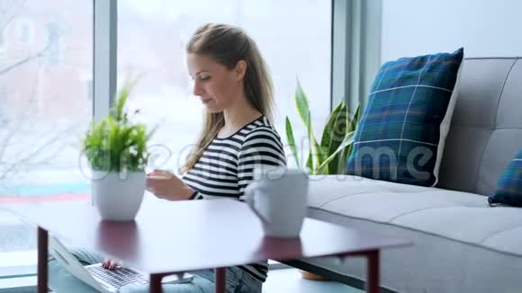 漂亮的年轻女人在家里用手提电脑工作时使用手机视频的预览图