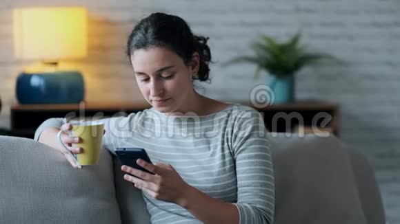 微笑的年轻女子使用她的手机而坐在沙发上的客厅在家里视频的预览图