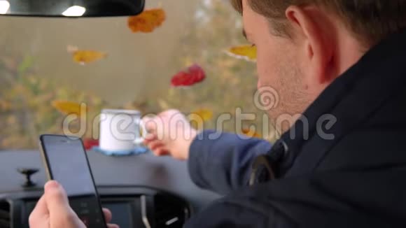 一个成年人坐在车里喝杯子里的茶用智能手机视频的预览图