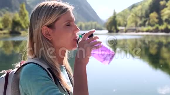 年轻漂亮的女旅行者背着背包站在湖边喝水视频的预览图