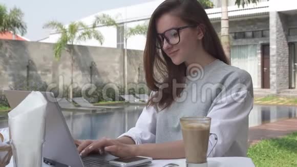 女人在笔记本电脑上工作在热带豪华别墅的游泳池附近带上智能手机计算机和移动方面的女性工作视频的预览图