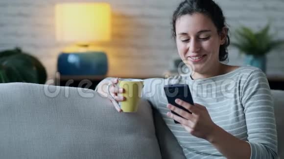 微笑的年轻女子使用她的手机而坐在沙发上的客厅在家里视频的预览图