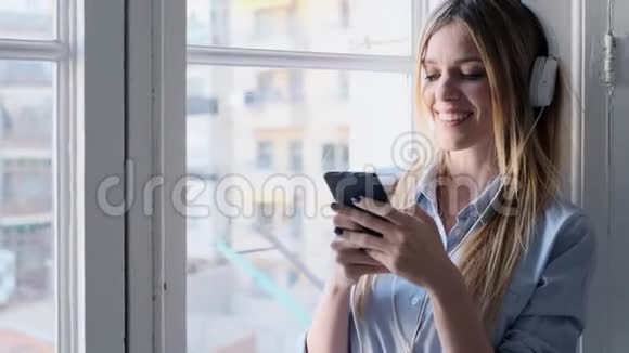 漂亮的年轻女人带着耳机听音乐在家里使用智能手机视频的预览图