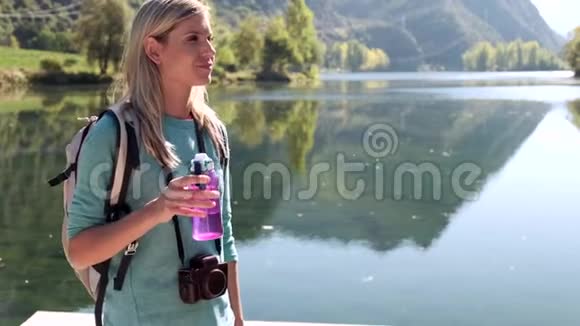 年轻漂亮的女旅行者背着背包站在湖边喝水视频的预览图