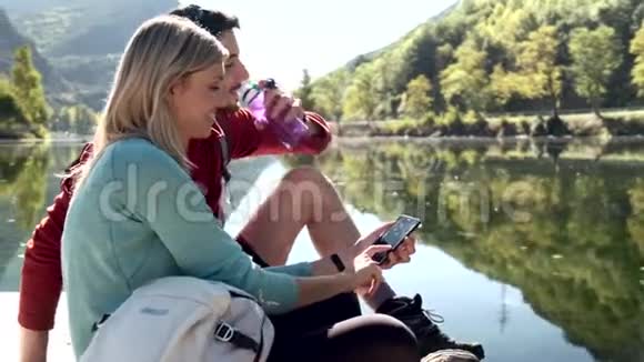 两个旅行徒步旅行者在山上坐在湖边时使用手机和饮用水视频的预览图