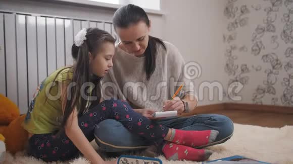 妈妈带着女儿坐在房间里毛茸茸的地毯上的地板上在一本小笔记本里画草图视频的预览图