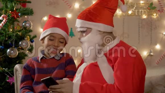 快乐的小男孩和圣诞老人坐在沙发上看着智能手机上的卡通视频的预览图