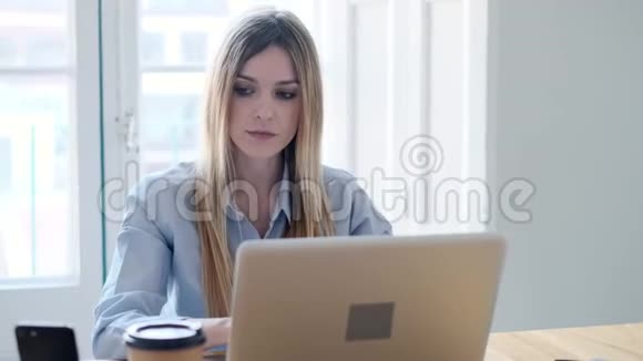 微笑的年轻商务女性使用她的智能手机同时在办公室工作笔记本电脑视频的预览图
