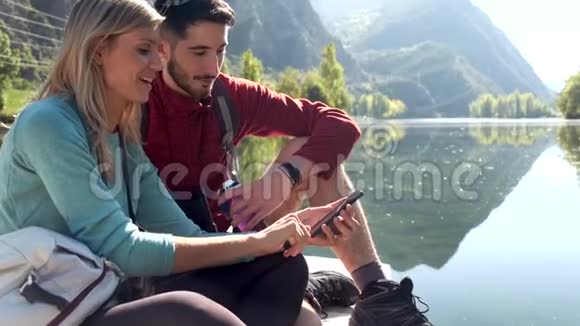 两名徒步旅行者在山上坐在湖边时使用手机视频的预览图