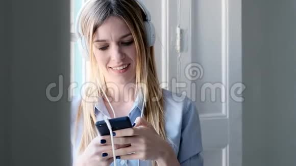 漂亮的年轻女人带着耳机听音乐在家里使用智能手机视频的预览图