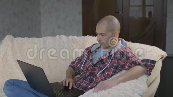 男性自由职业者坐在公寓的沙发上手里拿着一台笔记本电脑视频的预览图
