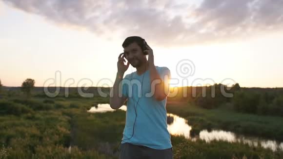 一个年轻人戴着大耳机听音乐在湖边日落时跳舞视频的预览图