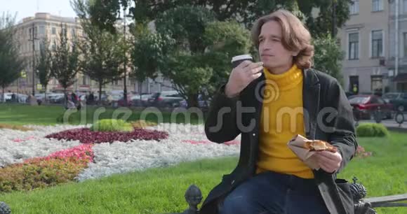 年轻人在公园里喝咖啡吃甜蛋糕他微笑着穿着一件黄色的毛衣和黑色的衣服视频的预览图