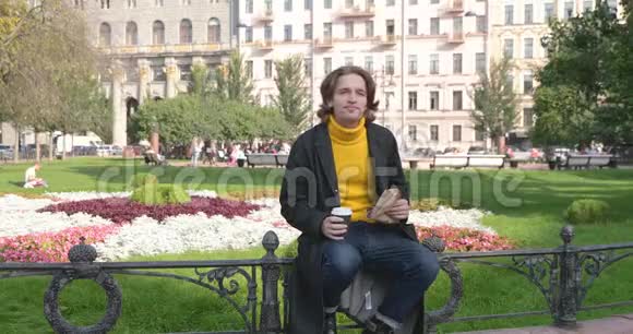 年轻人在公园里喝咖啡吃甜蛋糕他微笑着穿着一件黄色的毛衣和黑色的衣服视频的预览图
