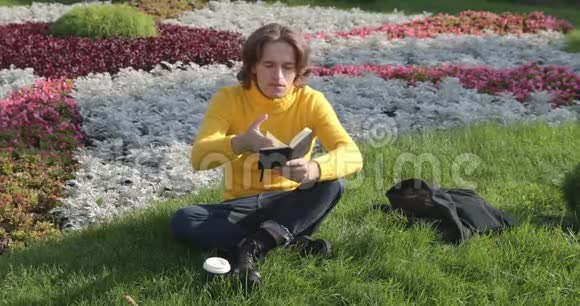 年轻人在公园里喝咖啡看书他微笑着休息一下穿着一件黄色毛衣视频的预览图