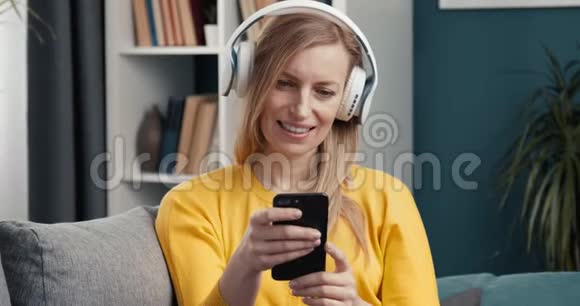 带着耳机和智能手机坐在沙发上的快乐女性视频的预览图