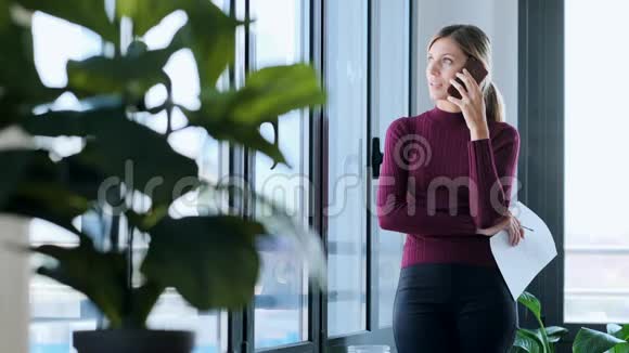 成功的年轻商务女性在办公室里站在窗口边工作一边用手机和阅读文件视频的预览图