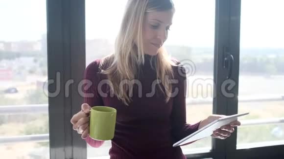 漂亮的年轻商务女性在办公室的窗户旁边坐着用她的数字平板电脑和喝咖啡视频的预览图