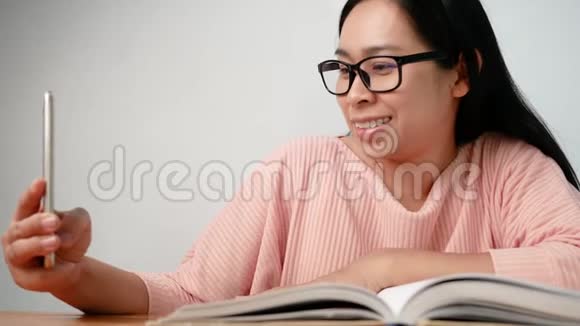 亚洲年轻女性使用智能手机坐在客厅里一边在家看书一边放松视频的预览图