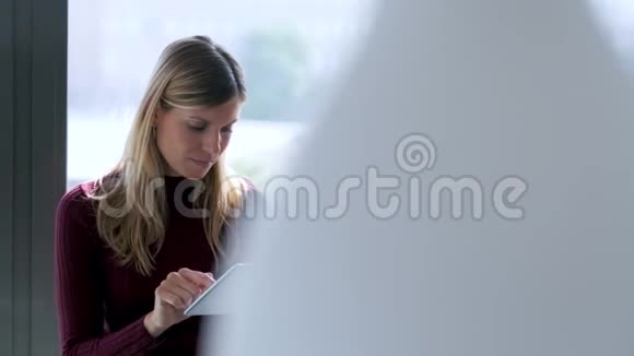 漂亮的年轻商务女性在办公室坐在窗户旁边时使用她的数字平板电脑视频的预览图