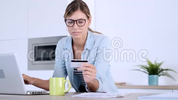 漂亮的年轻女人用她的笔记本电脑上网购物用信用卡付款而坐在厨房在家里视频的预览图