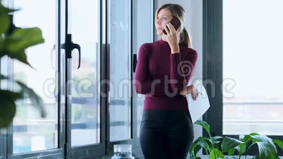 成功的年轻商务女性在工作中一边在办公室的窗户附近打电话一边看文件视频的预览图
