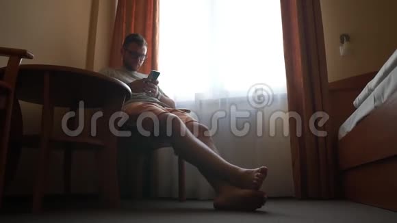 一个戴眼镜的男人坐在酒店房间窗户附近的椅子上拿着电话视频的预览图