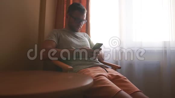 一个男人坐在酒店房间窗户附近的椅子上用电话工作视频的预览图