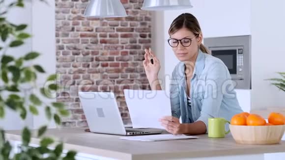 漂亮的年轻女人在家里厨房里用笔记本电脑和文件工作视频的预览图