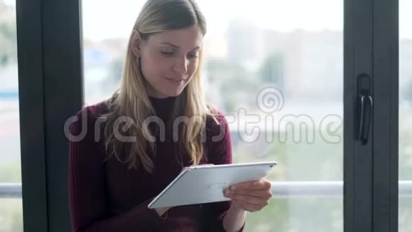 漂亮的年轻商务女性在办公室坐在窗户旁边时使用她的数字平板电脑视频的预览图