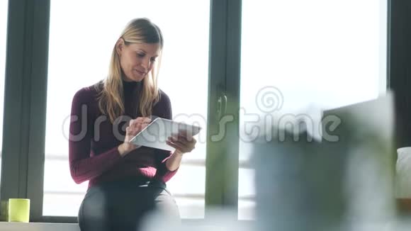 漂亮的年轻商务女性坐在办公室的窗户旁边一边看着相机一边用她的数字平板电脑视频的预览图