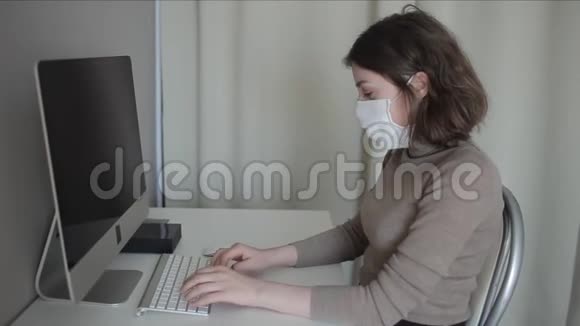 在隔离期间一个年轻女孩戴着医疗面罩在家里的电脑上工作冠状病毒COVID19视频的预览图