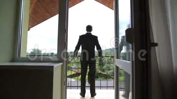 英俊自信的新郎穿着黑色西装站在阳台上靠在栏杆上看着绿色的森林视频的预览图