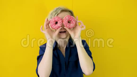 疯狂开朗的金发女孩微笑着玩得很开心眼睛上看着两个红色甜甜圈糖果黄色工作室视频的预览图